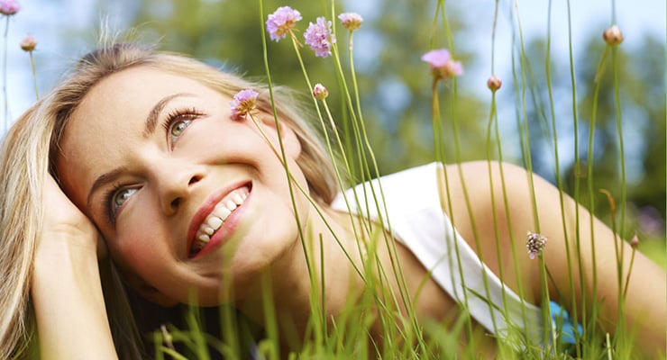 Mujer recostada sobre el césped sonriendo entre flores pequeñas al aire libre.