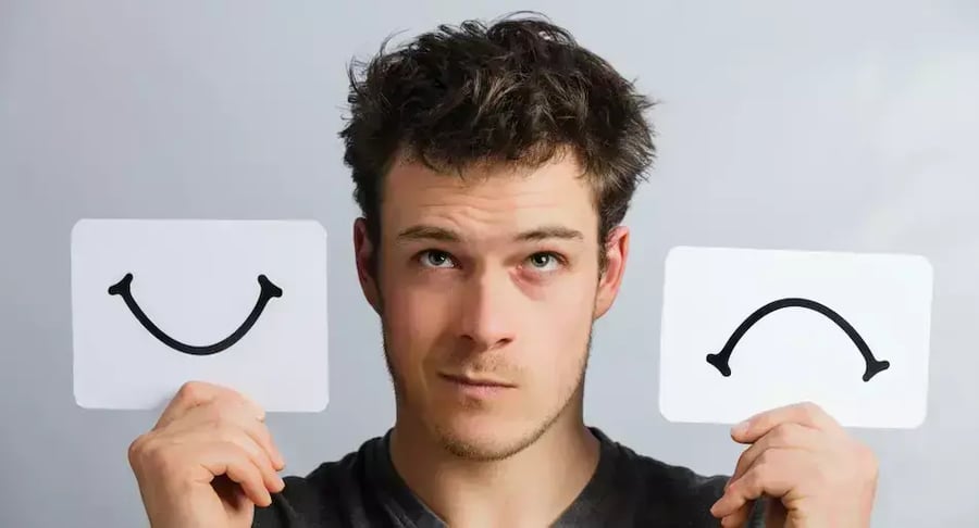 Hombre joven sosteniendo tarjetas con caritas feliz y triste como símbolo de salud mental