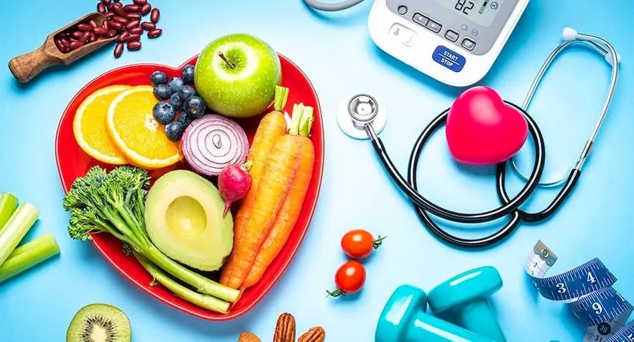 Alimentos saludables dispuestos en un plato con forma de corazón junto a un estetoscopio, tensiómetro y pesas, sobre un fondo azul.
