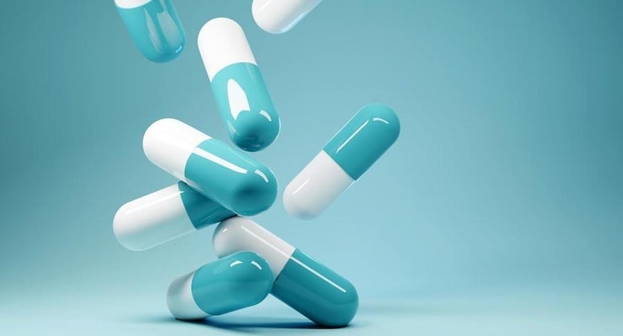 Varias cápsulas médicas de color blanco y celeste cayendo o flotando sobre un fondo azul claro uniforme, representando el suministro de medicamentos.