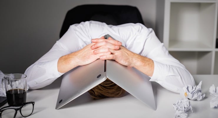 Hombre de negocios con camisa blanca recostado sobre su escritorio con la cabeza cubierta por su computadora portátil, rodeado de papeles arrugados, representando agotamiento laboral.
