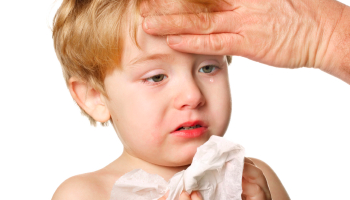 Niño pequeño con malestar y congestión nasal mientras un adulto le toma la temperatura con la mano para revisar si tiene fiebre.
