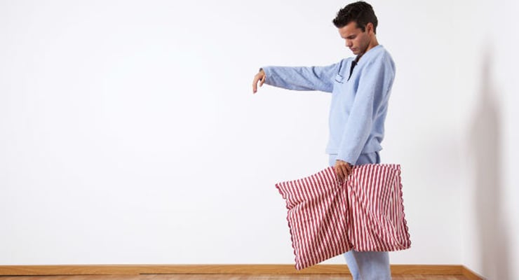Hombre caminando dormido en pijama sosteniendo una almohada dentro de una habitación