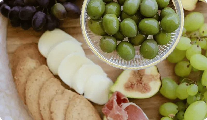 Tazón de aceitunas verdes junto a uvas, higos, galletas y rodajas de queso sobre una tabla.