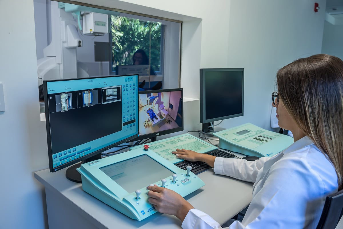 Especialista médica con bata blanca operando una consola de control y monitores digitales de rayos X en una sala de diagnóstico radiológico.