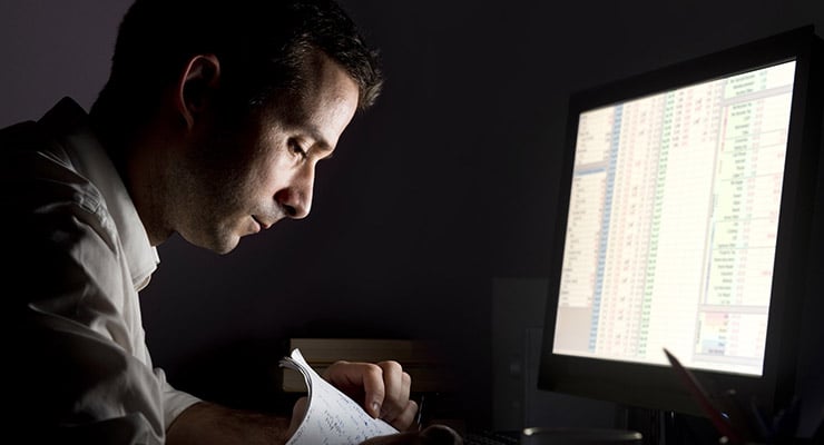 Hombre trabajando de noche frente a una computadora revisando documentos impresos.