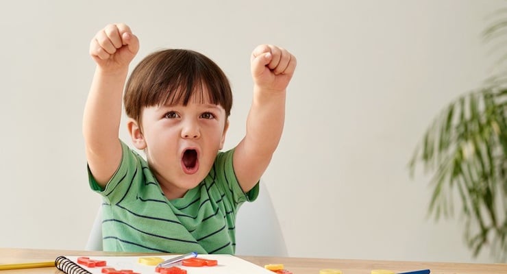 Un niño pequeño con camisa verde grita con entusiasmo y levanta los puños mientras realiza tareas escolares, representando la impulsividad o hiperactividad.