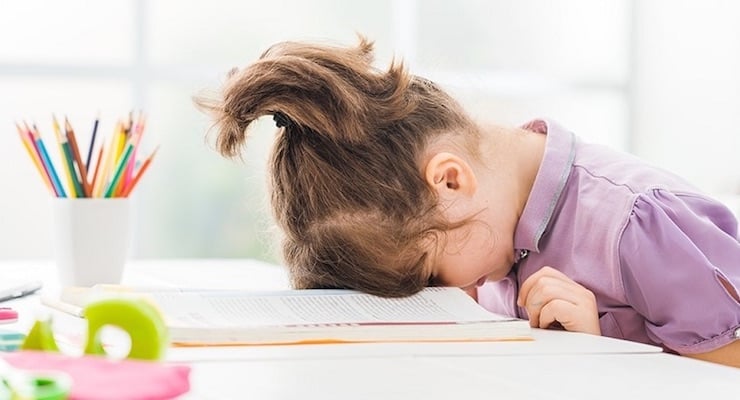 Una niña pequeña con el cabello recogido apoya su cabeza sobre un libro abierto en un escritorio, mostrando señales de frustración o cansancio durante el estudio.