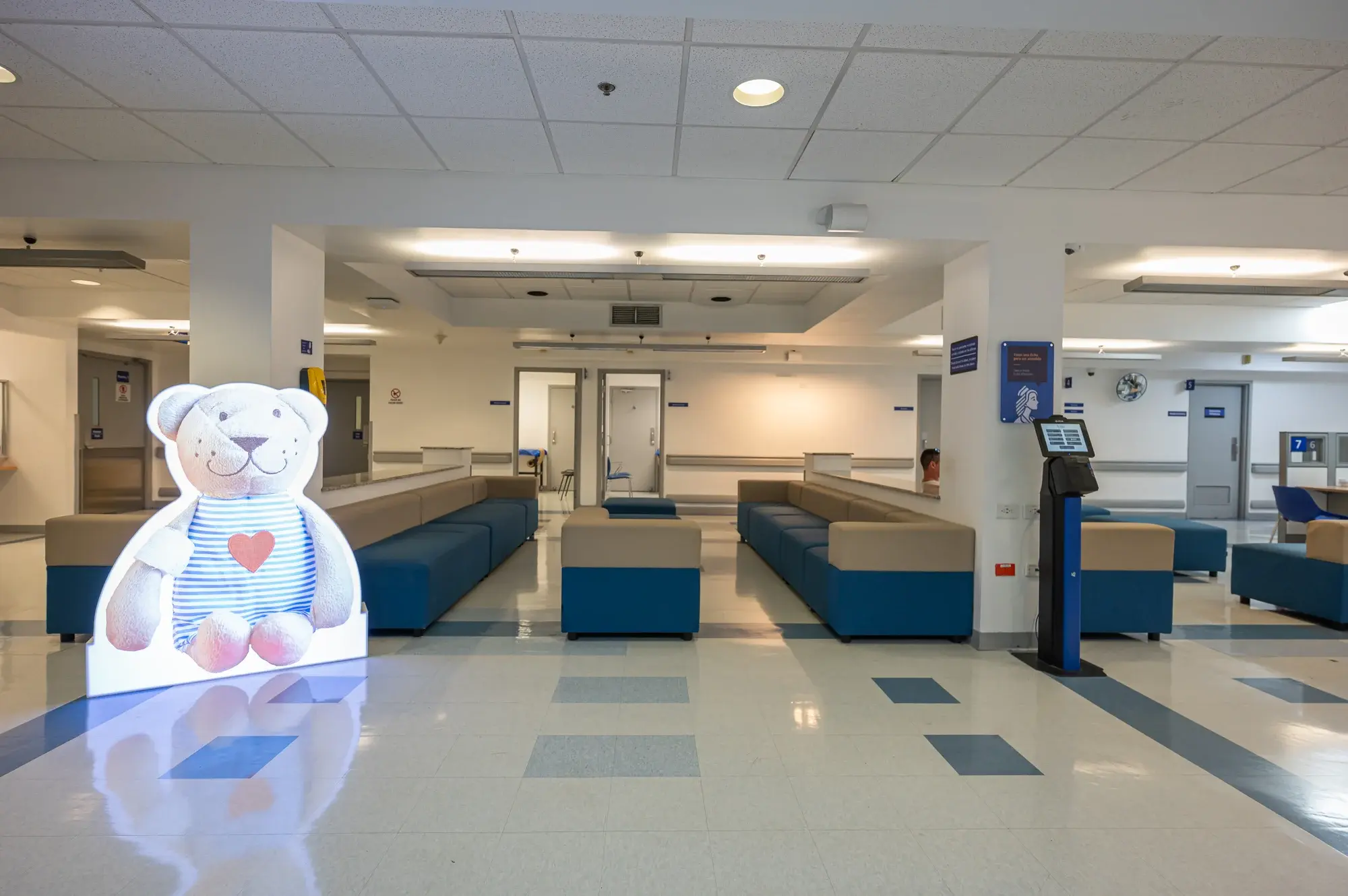 Sala de espera de hospital con sillones azules y beige, decoración infantil con oso de peluche.