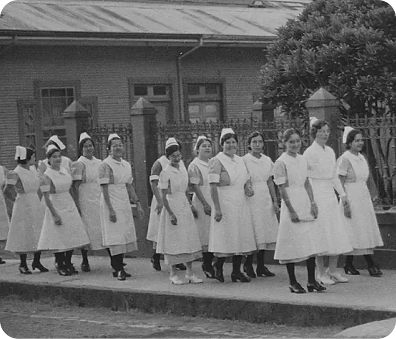 Grupo de enfermeras con uniforme blanco desfilando frente a un edificio antiguo en fotografía en blanco y negro.