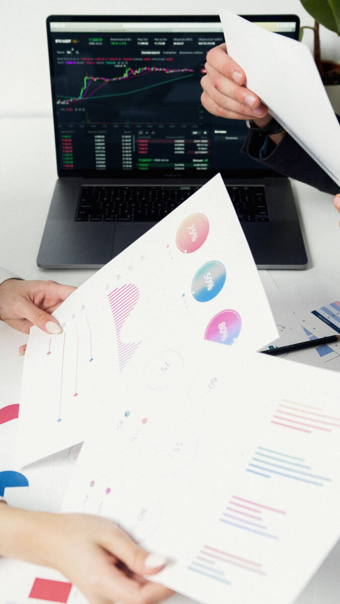 Un primer plano elevado de dos pares de manos y una laptop analizando gráficos financieros complejos en una pantalla interactiva y en papel en un entorno de innovación.