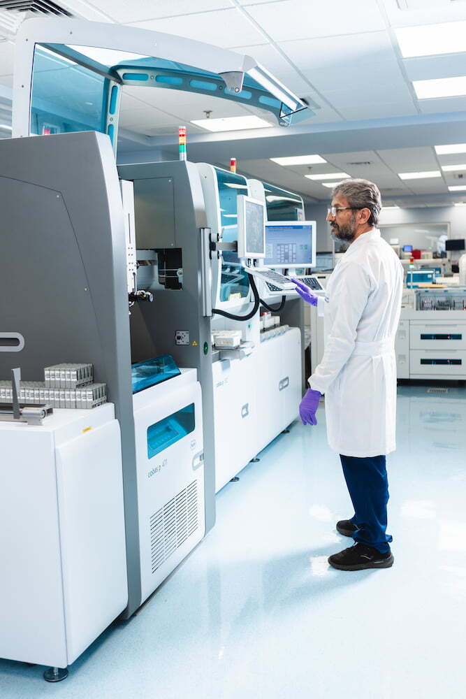 Especialista con bata blanca y guantes morados operando un moderno analizador automatizado en un laboratorio clínico.