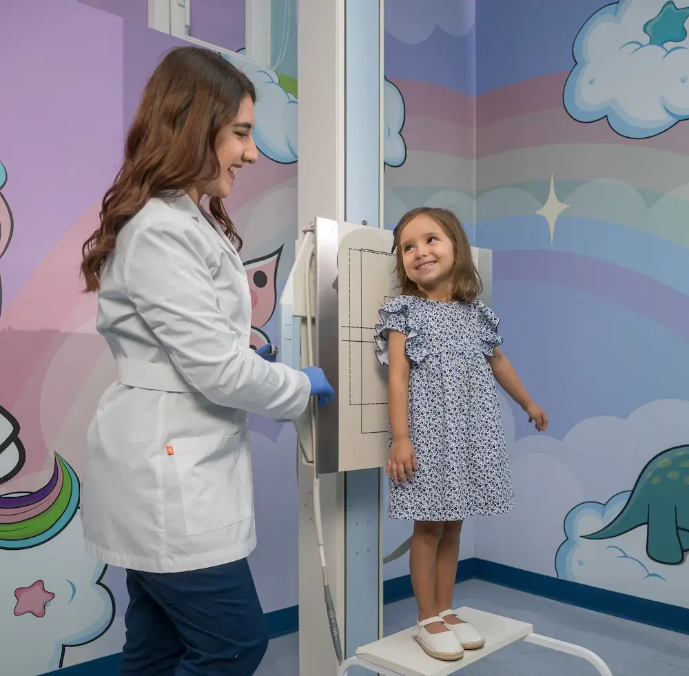 Especialista en radiología del Hospital Clínica Bíblica atendiendo de forma empática a una niña para un examen de rayos X en una sala con decoración infantil.