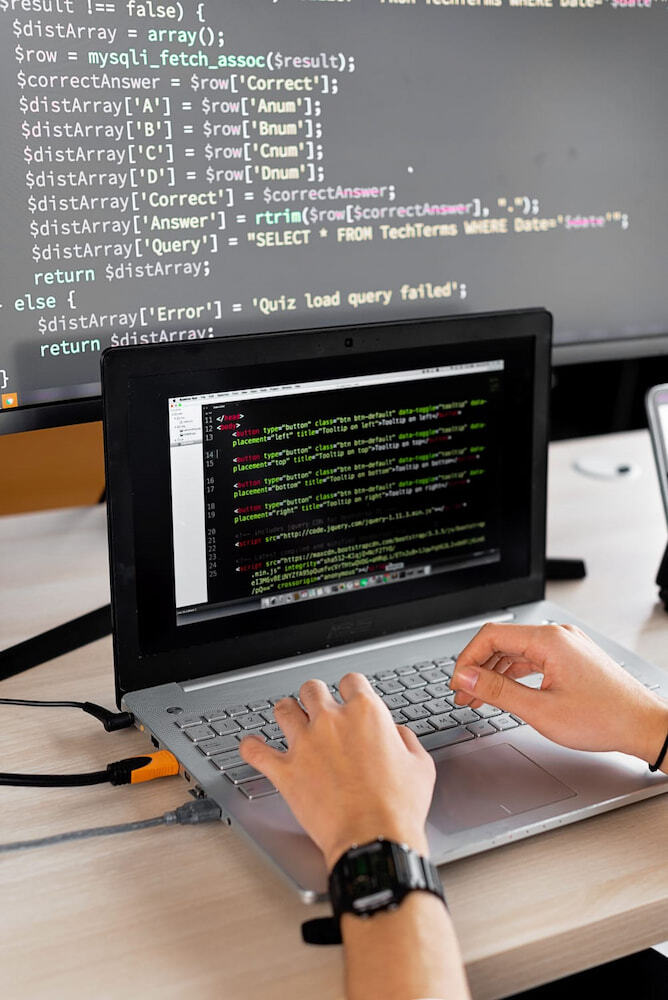 Manos de un programador escribiendo líneas de código en una laptop con un monitor secundario al fondo.