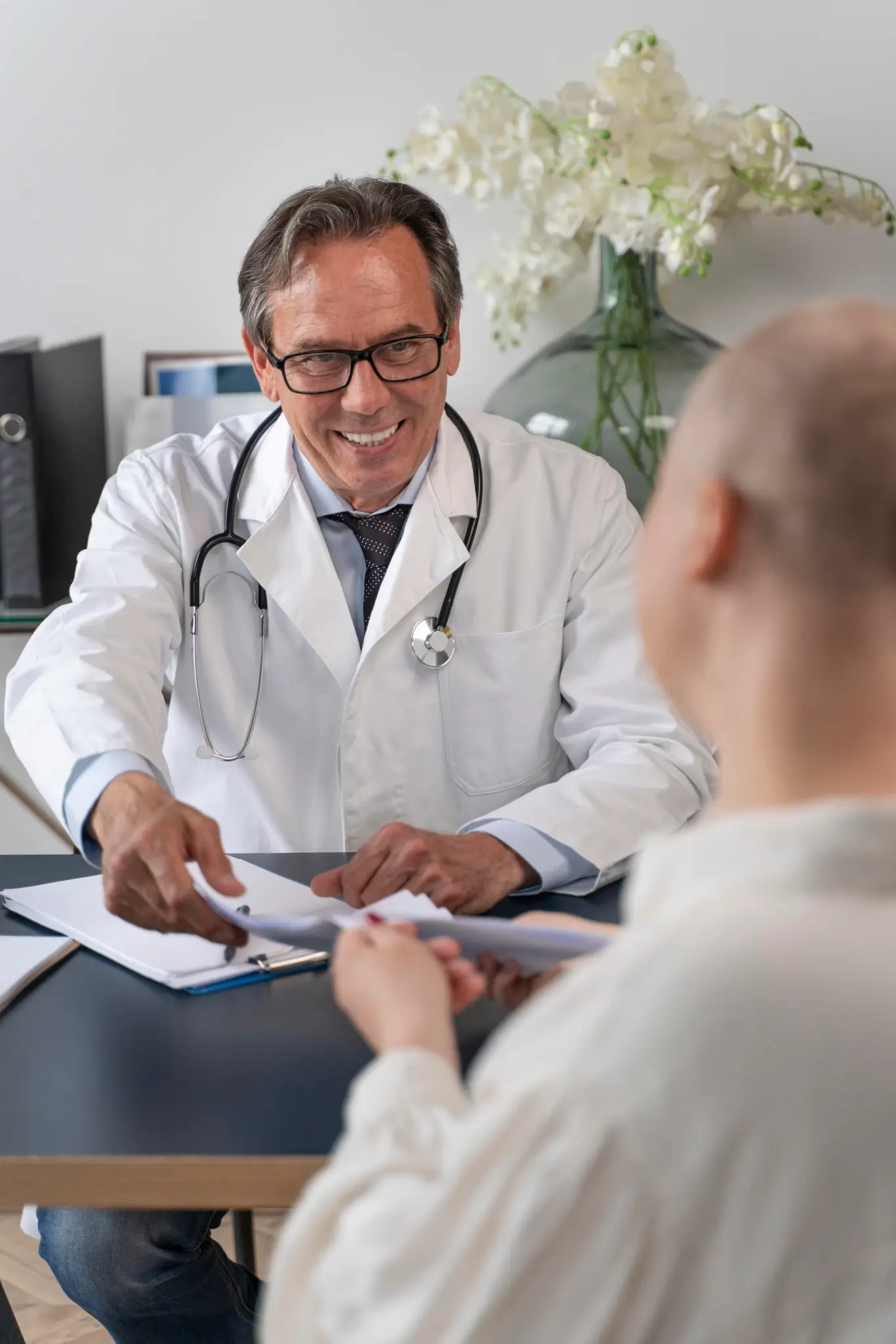 Un doctor con bata blanca y estetoscopio sonríe mientras entrega unos resultados médicos o documentos a una paciente en su consultorio.