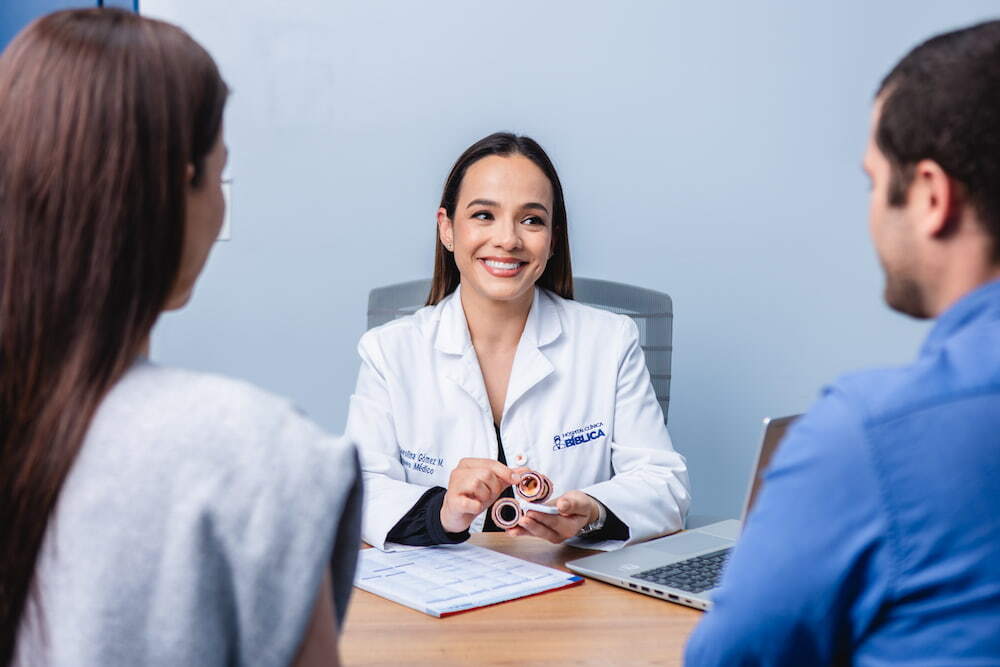 Doctora sonriente con bata blanca sentada en su escritorio explicando un modelo anatómico a una pareja de pacientes de espaldas en un consultorio.