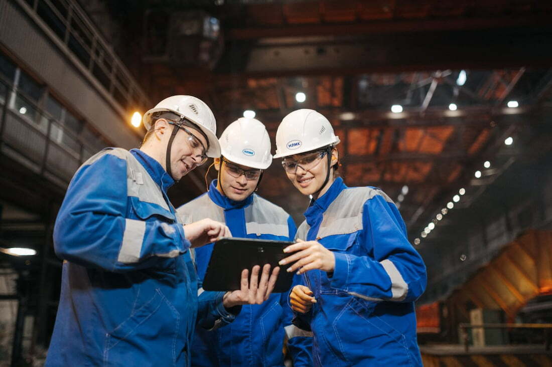 Tres ingenieros con casco y uniforme azul colaboran revisando una tableta digital dentro de una planta industrial.