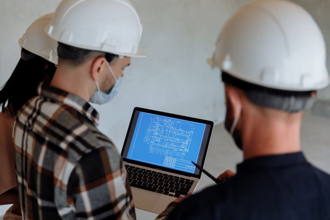 Tres profesionales con cascos de seguridad revisan un plano arquitectónico digital en una laptop.