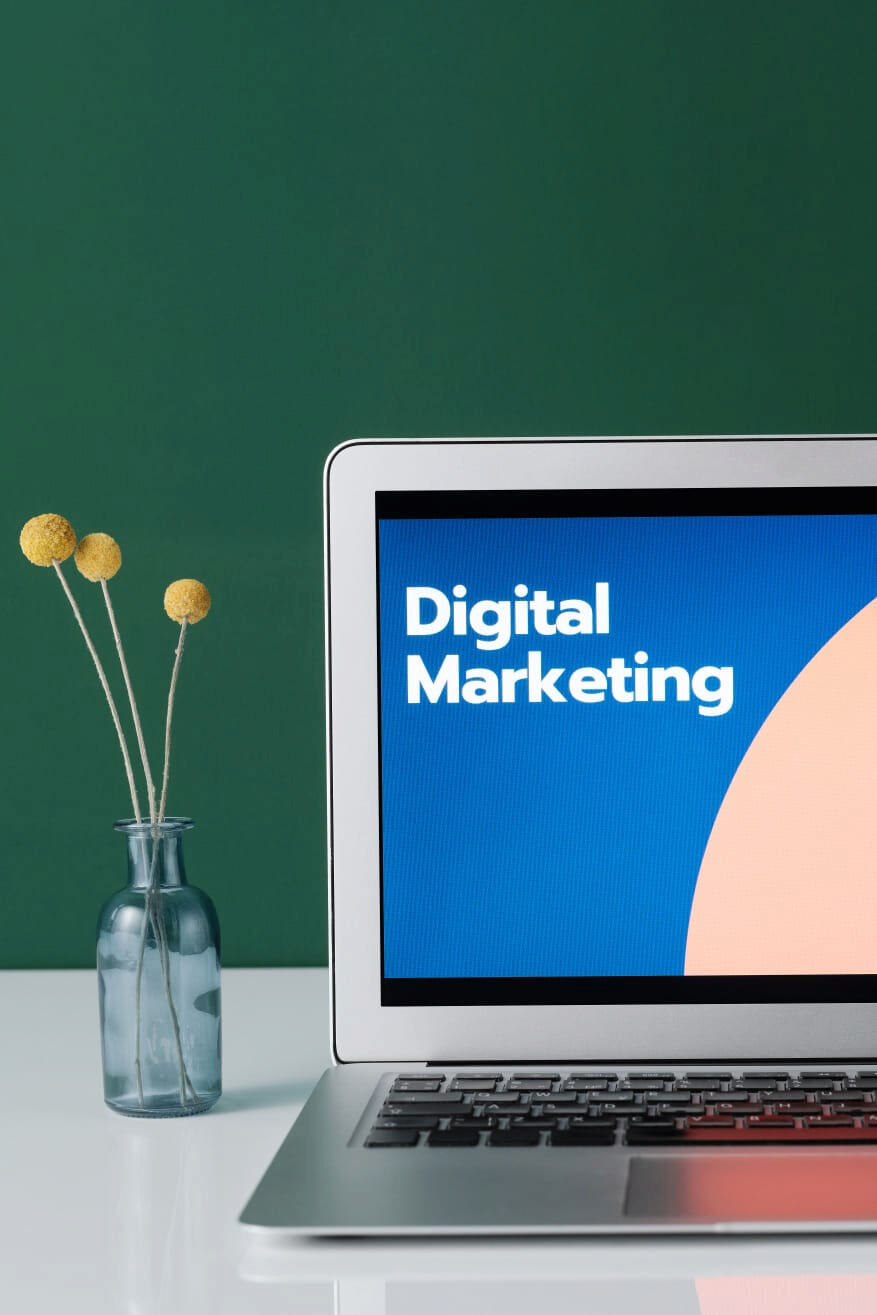 Una laptop abierta que muestra en su pantalla el texto Digital Marketing junto a un pequeño florero sobre un fondo verde.