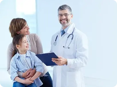 Un médico sonriente con bata blanca y estetoscopio sostiene una carpeta clínica mientras conversa de manera empática con una madre y su hija pequeña.