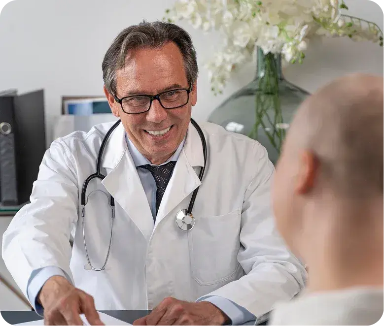 Un médico especialista sonriente con anteojos y estetoscopio, mirando a un paciente cuyo rostro está en la sombra, durante una consulta médica empática en el Hospital Clínica Bíblica.
