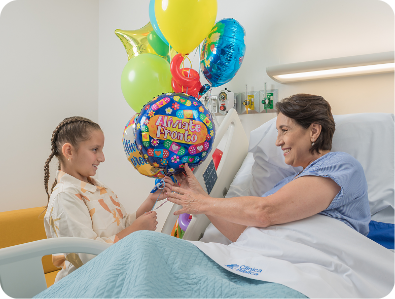 Niña entregando globos de colores con mensaje “Alíviate pronto” a una paciente sonriente en cama de hospital.