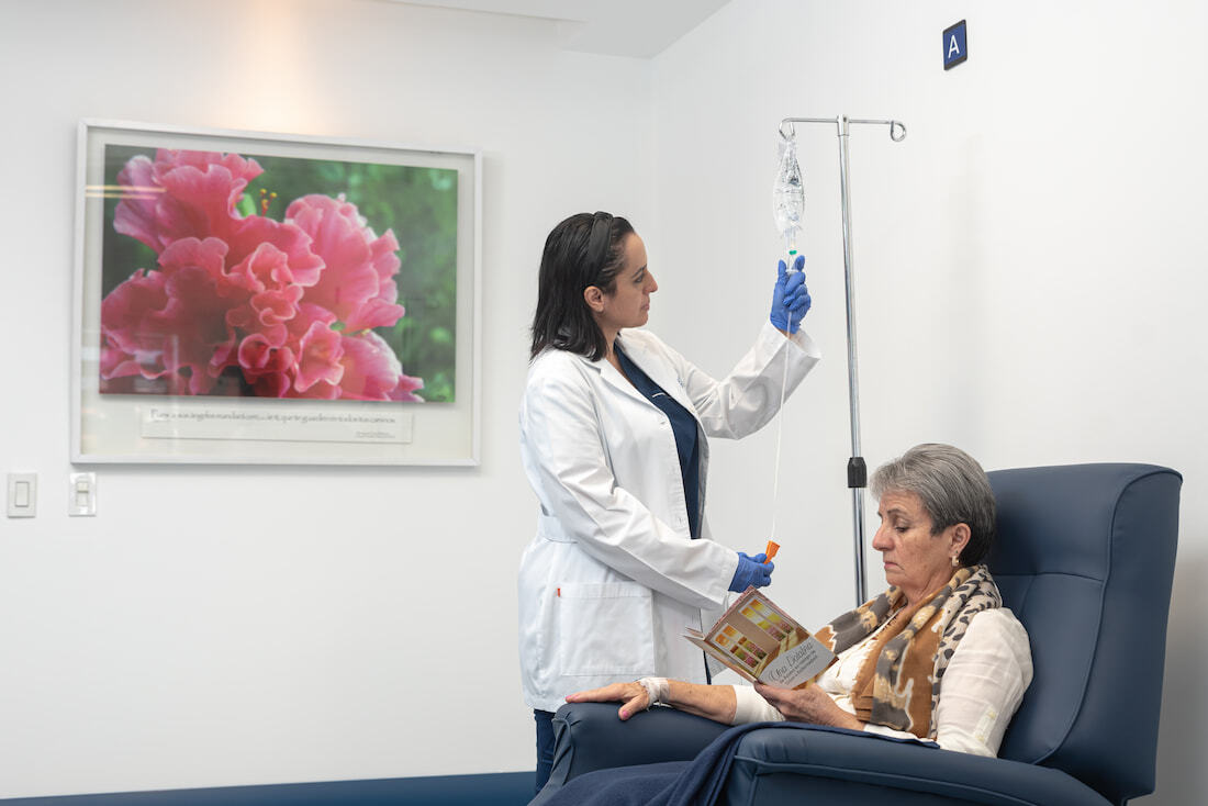 Doctora con bata blanca ajustando una solución intravenosa a una paciente mayor sentada en un sillón médico dentro de una clínica.