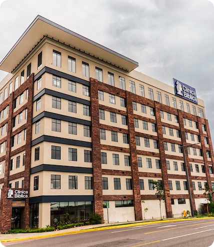 Fachada del edificio principal de la Clínica Bíblica con letrero visible en la parte superior y cielo nublado.